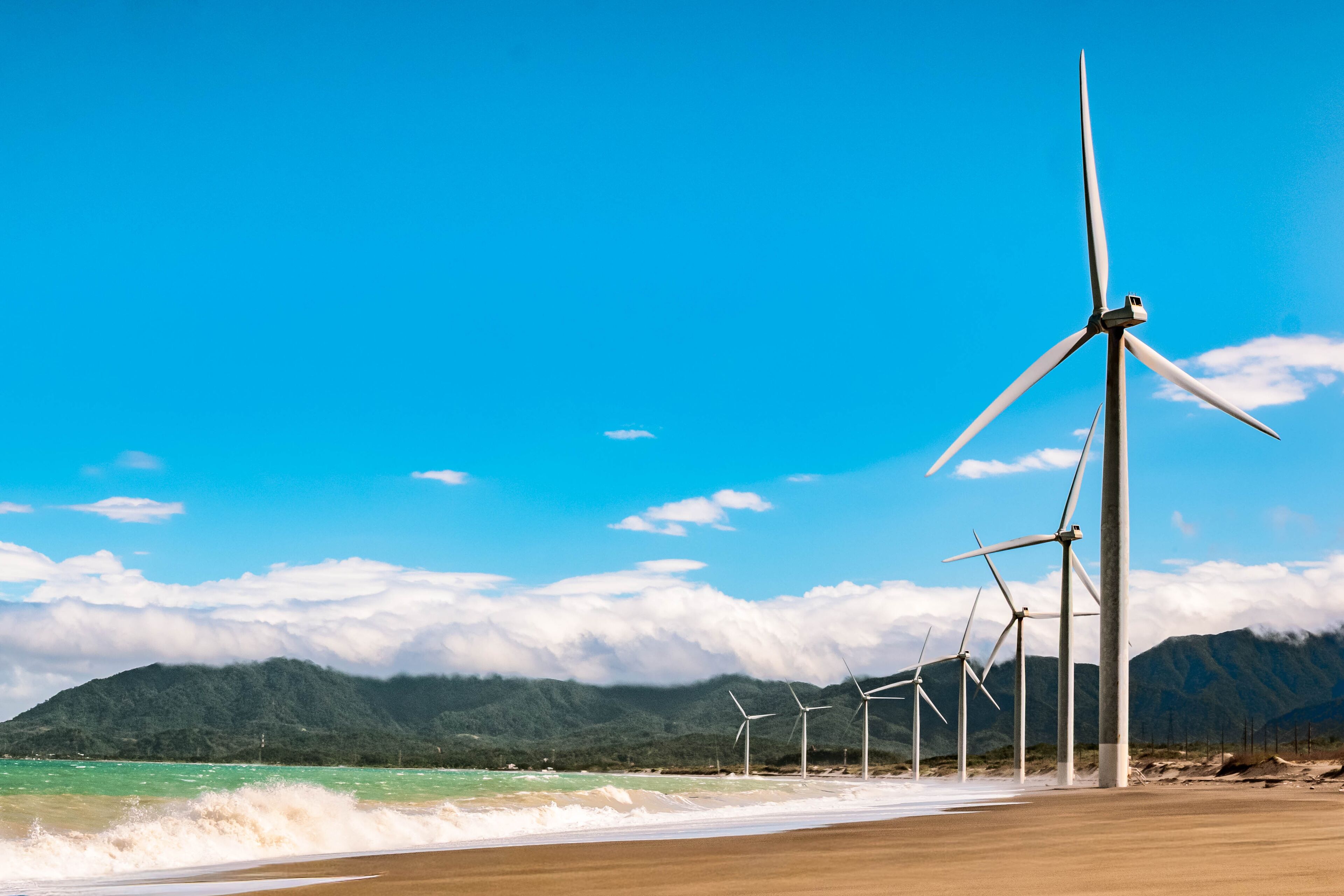 A beach with wind turbines and mountains in the background.