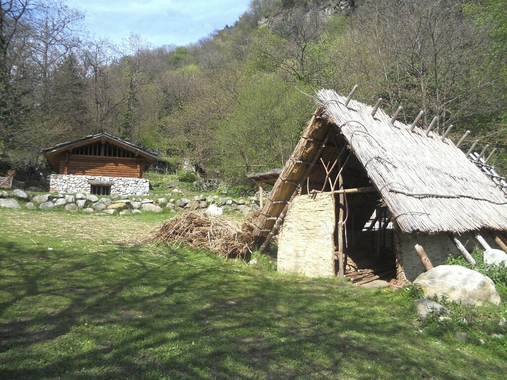 Una cabaña tradicional con techo de paja y base de piedra, rodeada de césped y un muro de piedra.