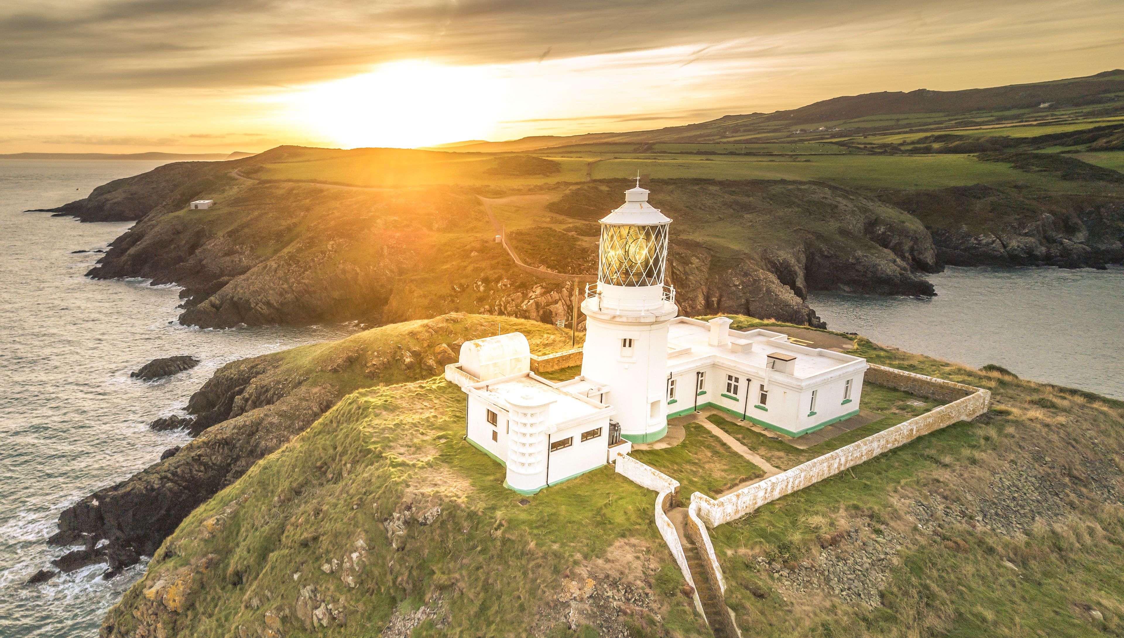 Un faro su una scogliera erbosa affacciata sul mare, con un tramonto sullo sfondo.