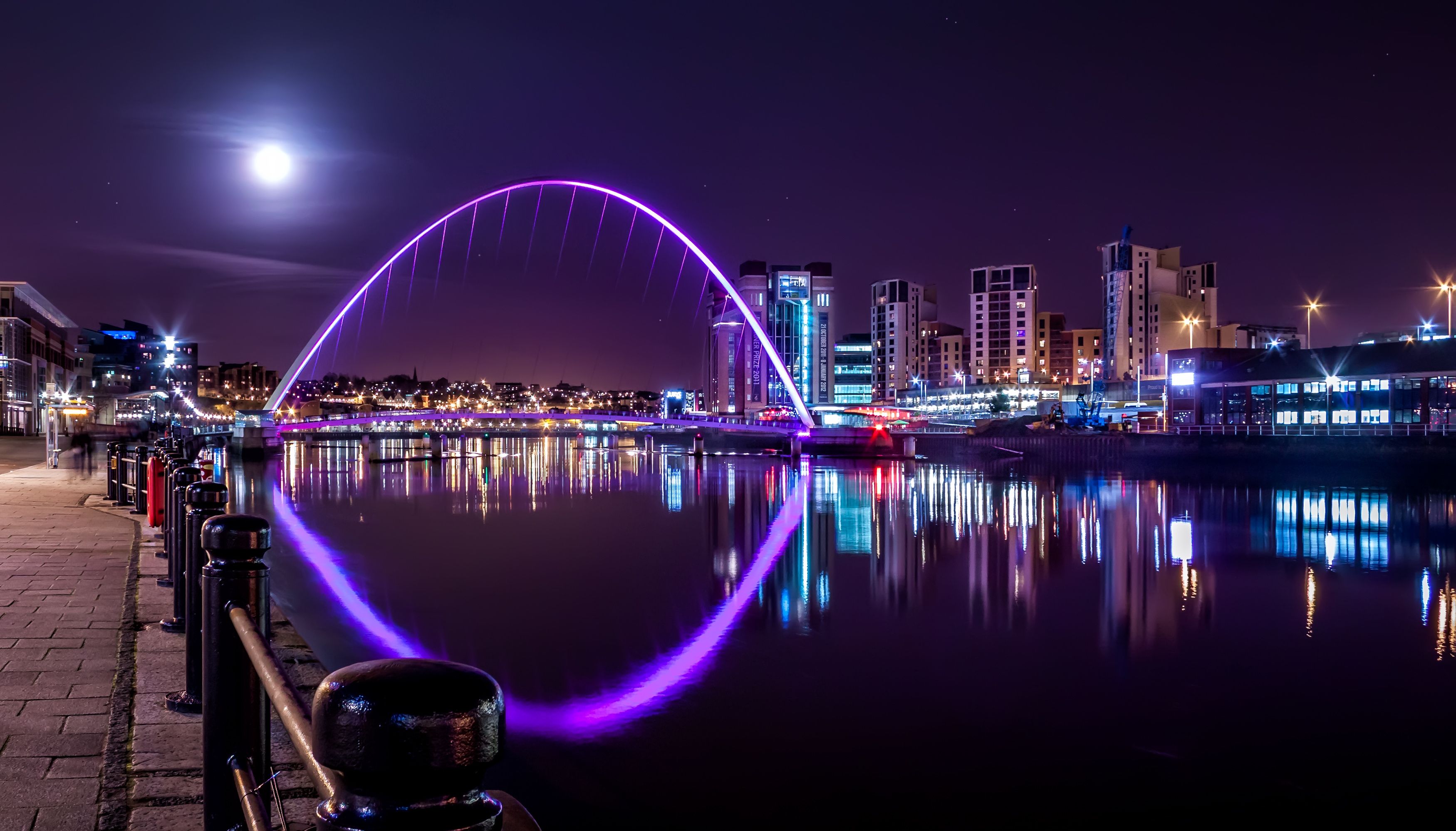 A cityscape at night with a brightly lit bridge reflecting on the water, illuminated buildings, and a clear sky.
