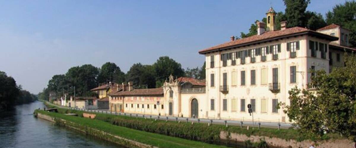 A canal with a row of classical buildings, a grassy embankment, and a clear sky.