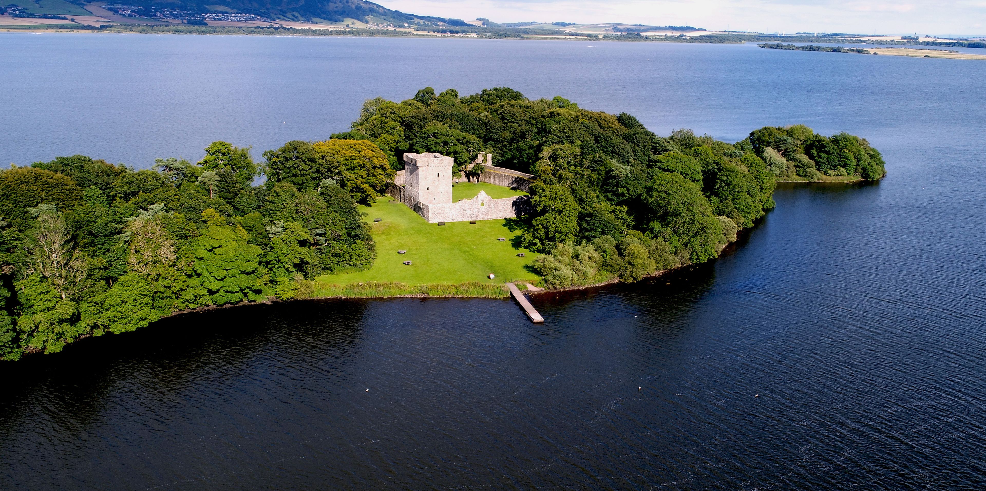 An aerial view of a small island with a castle surrounded by dense greenery and a calm body of water.