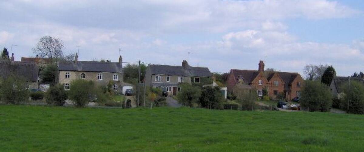A rural landscape with houses, a green field, and a partly cloudy sky.