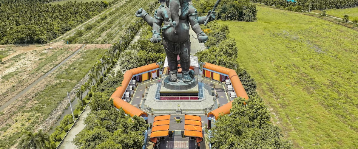 A large statue of a deity with multiple arms, surrounded by a circular platform and orange railings, set against a backdrop of green fields and distant power lines.