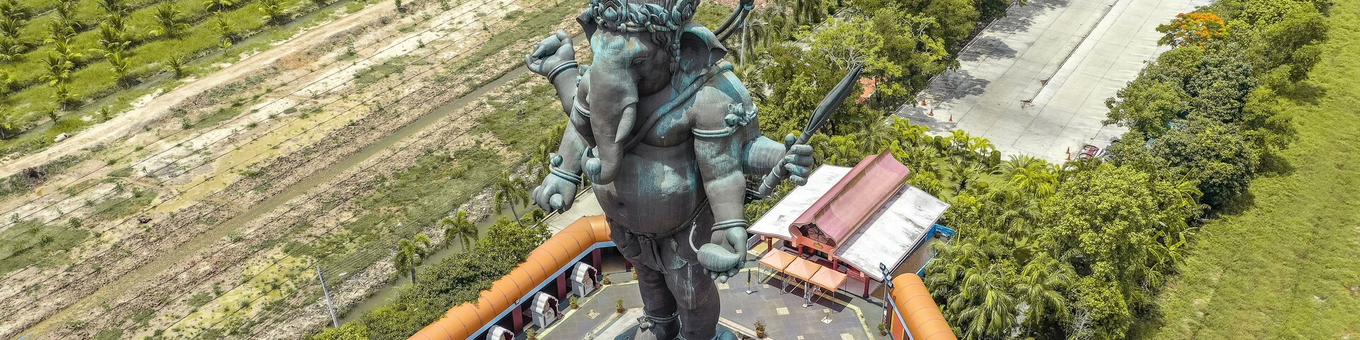 Una gran estatua de bronce de una deidad, rodeada por una plataforma circular, ubicada en un área ajardinada con palmeras y una carretera cerca.