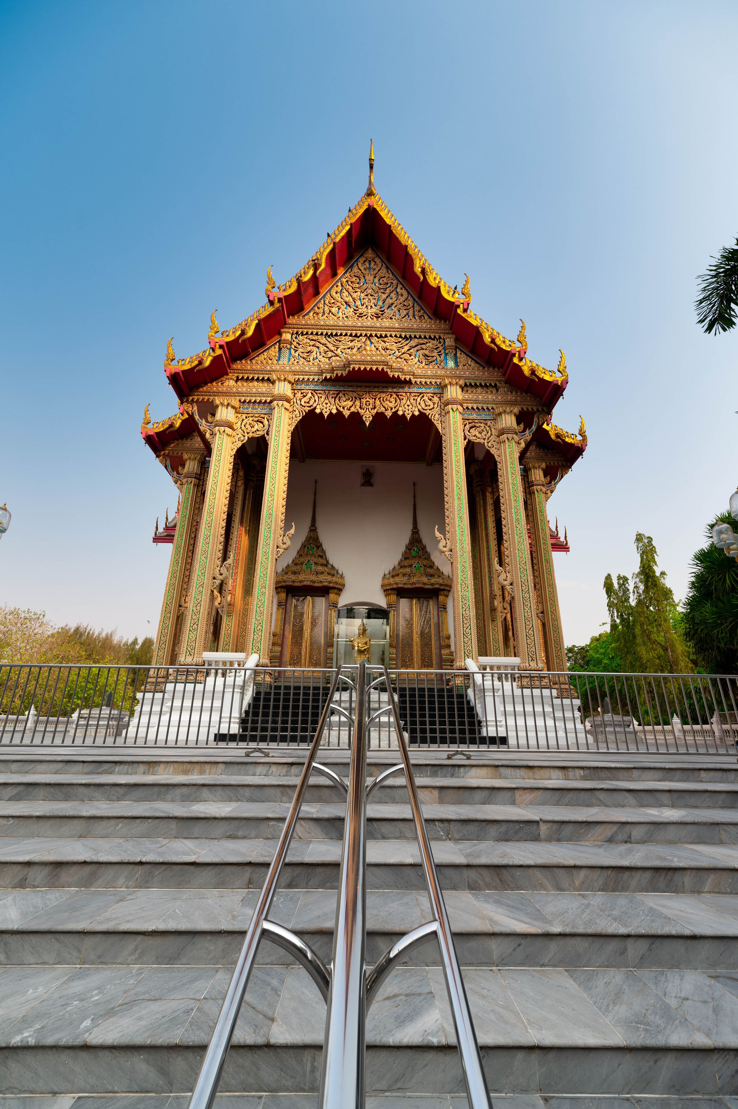 A temple with ornate golden decorations and a red roof, surrounded by stone steps and a metal railing.