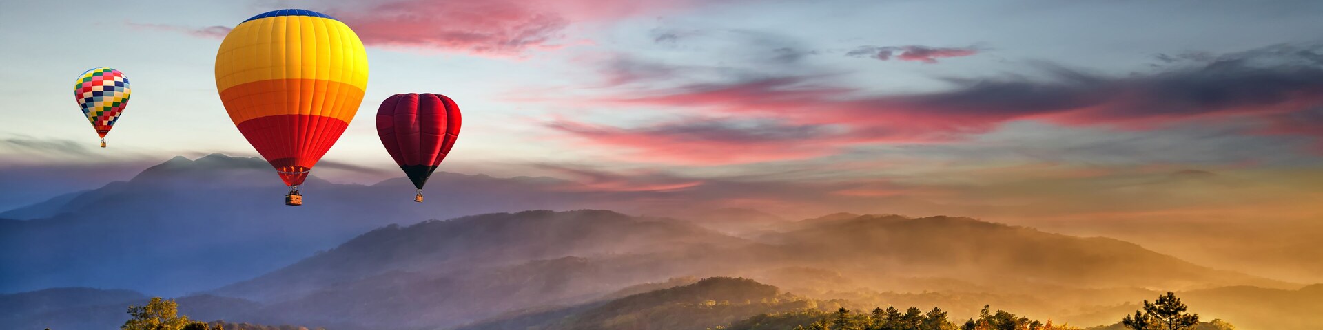Tres globos aerostáticos sobre un paisaje montañoso con amanecer o atardecer.