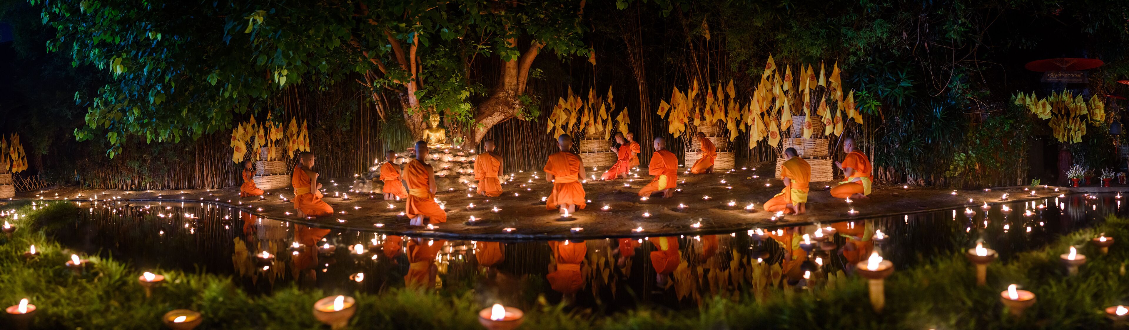 A serene nighttime scene with monks in orange robes around a pond, illuminated by candles and surrounded by trees and yellow flags.