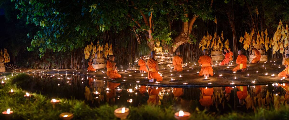 A serene nighttime scene with monks in orange robes around a pond, illuminated by candles and surrounded by trees and yellow flags.