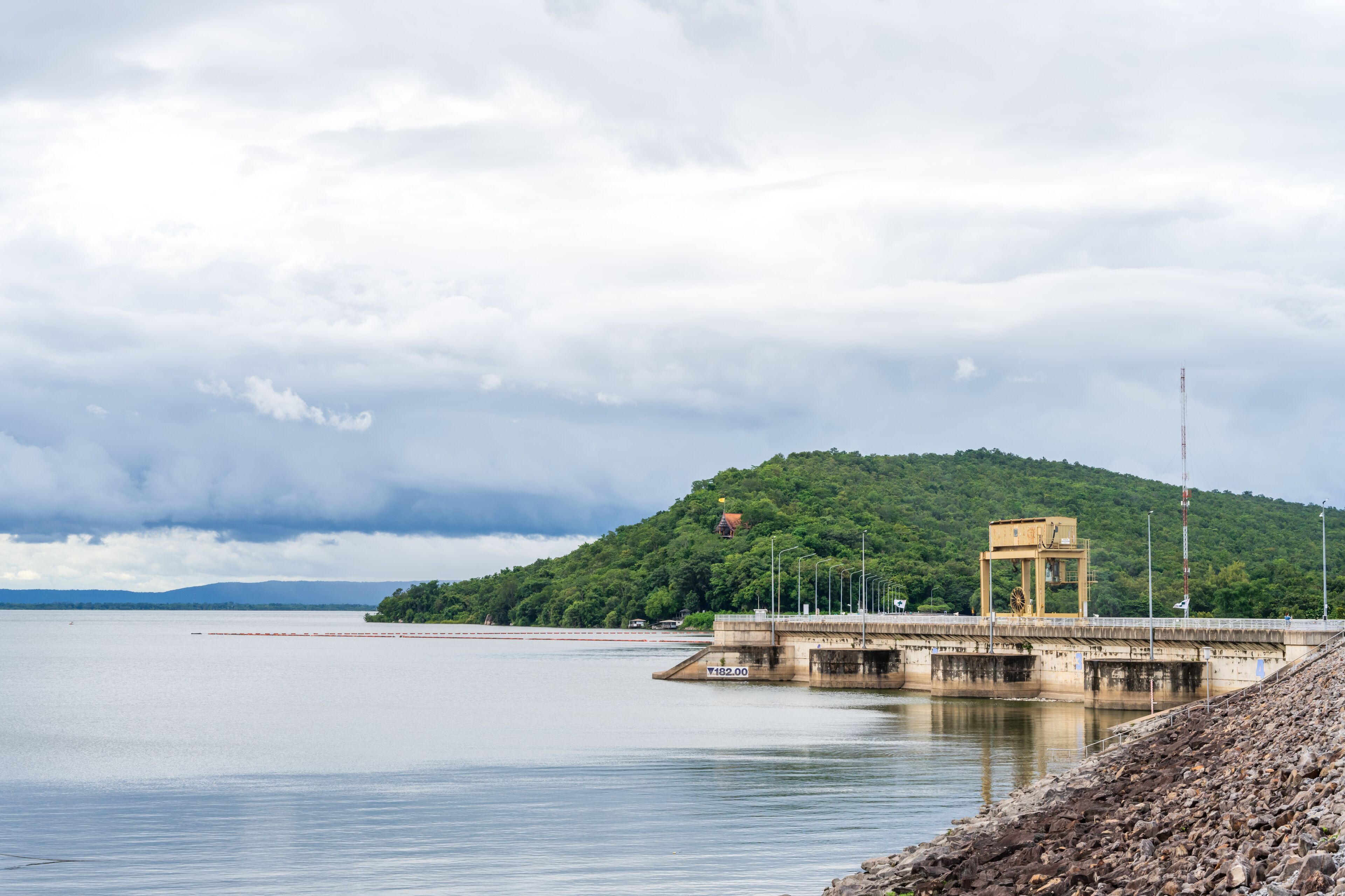 A dam with a control tower, calm water, and a forested hill in the background.