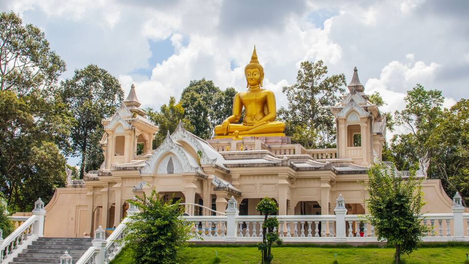 A large golden Buddha statue in front of a temple with a white fence and stairs.
