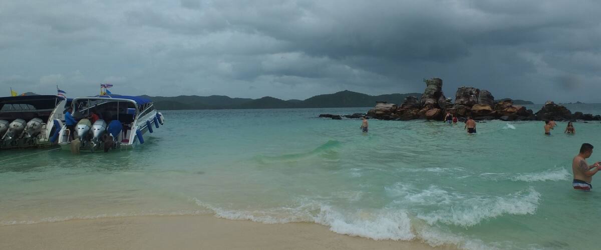 A beach with clear turquoise water, a boat with multiple outboard motors, and rocky formations in the distance.