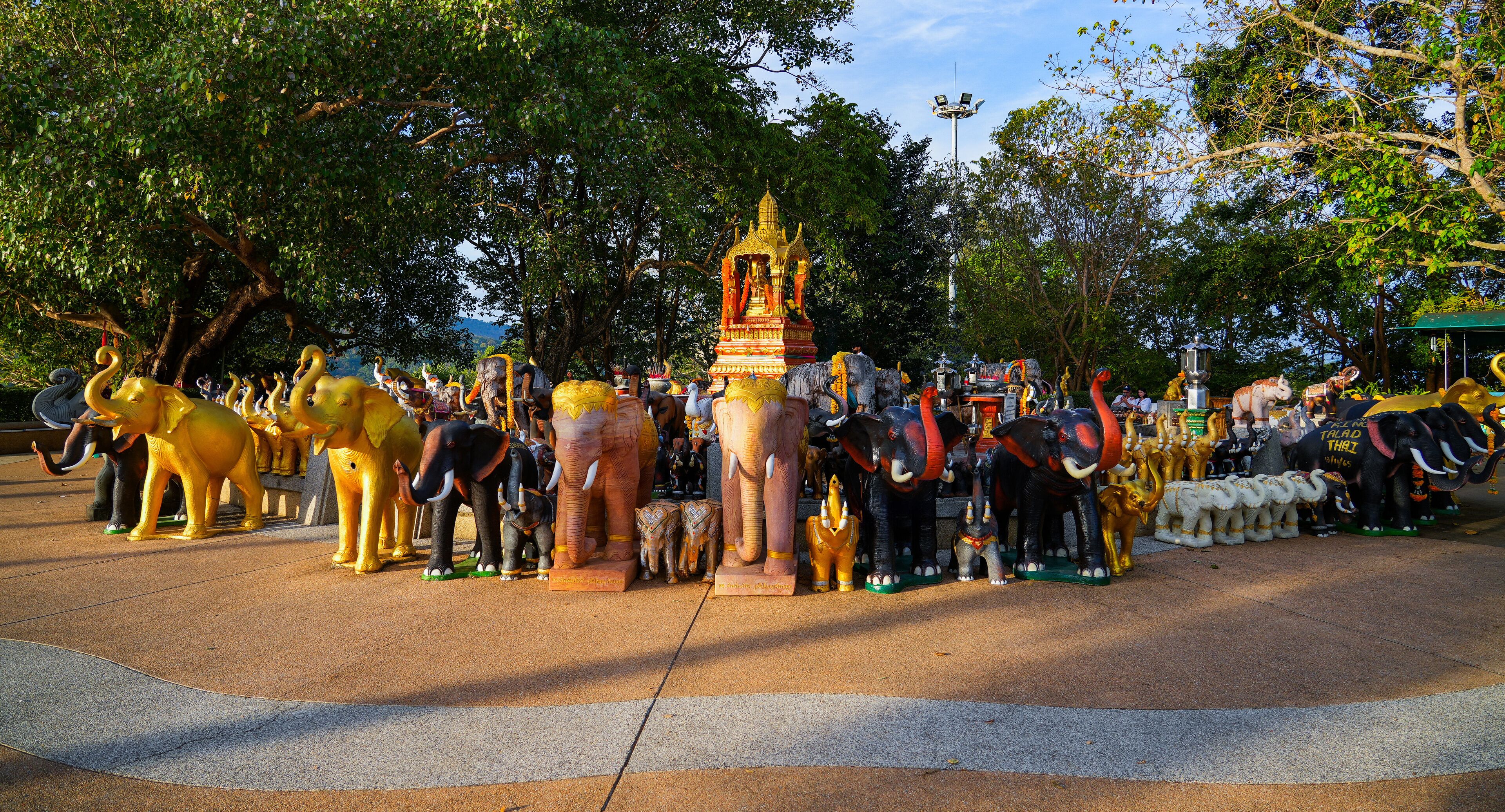 A collection of elephant statues in various colors and sizes, surrounded by trees and a temple-like structure.