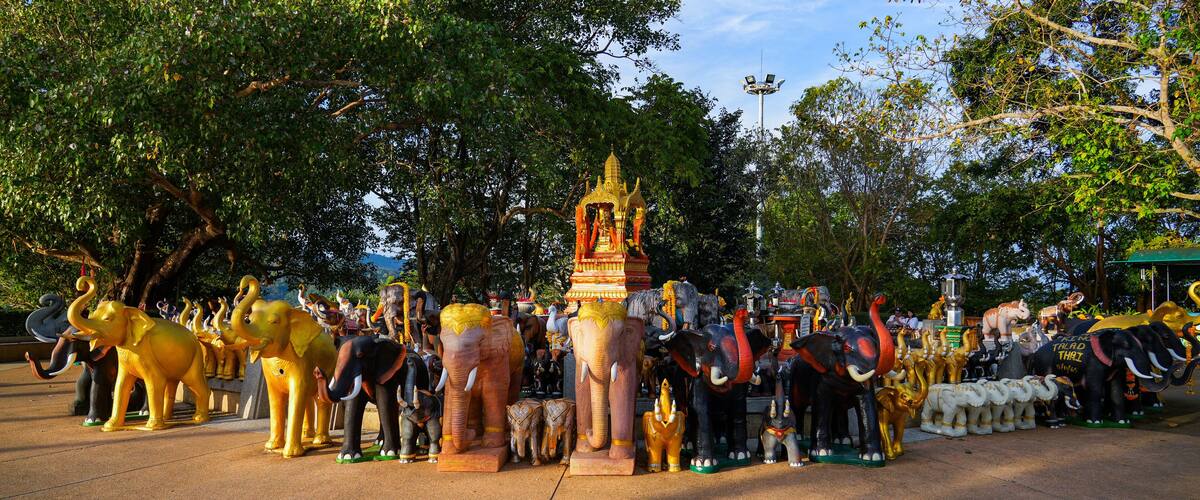 A collection of elephant statues in various colors and sizes, surrounded by trees and a temple-like structure.
