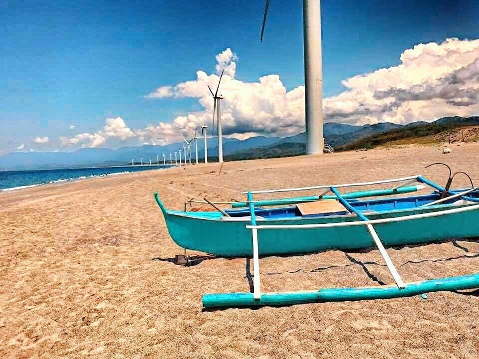 A beach with a row of wind turbines in the background and a blue boat on the sand.