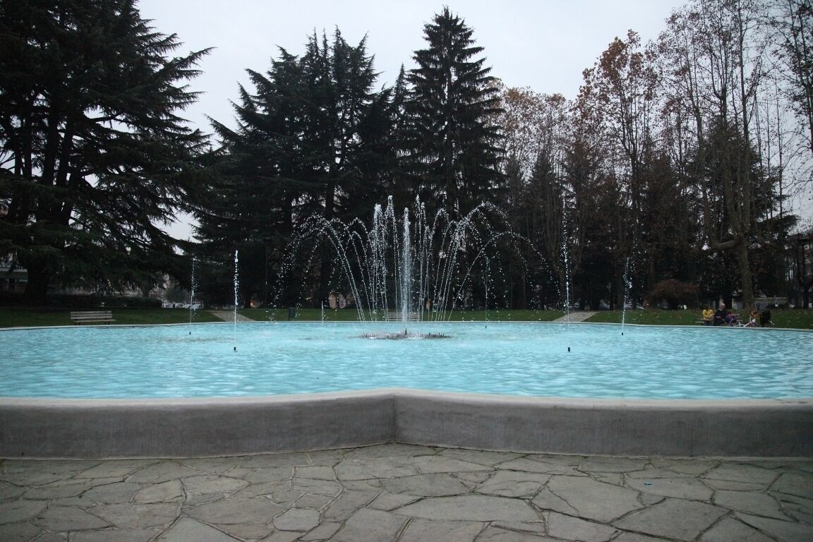 A fountain with water jets in a park with trees and a stone-paved area.