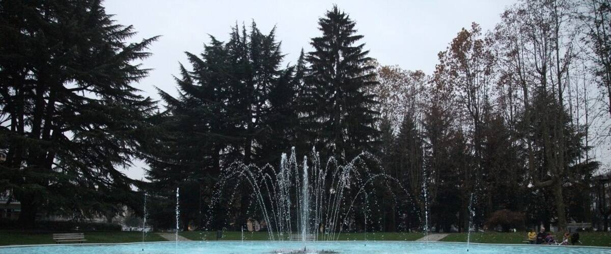 A fountain with water jets in a park with trees and a stone-paved area.