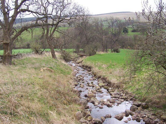 A rocky stream in a rural landscape with trees and hills in the background.