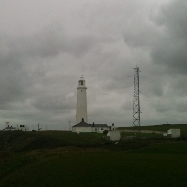 A lighthouse, a white tower, and a metal tower on a grassy hill.