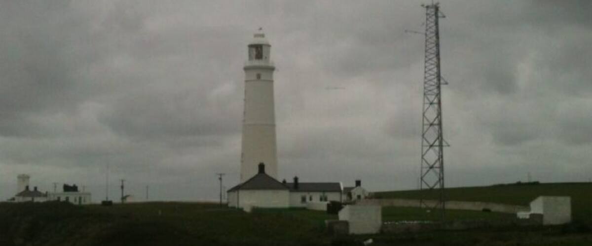 A lighthouse, a white tower, and a metal tower on a grassy hill.