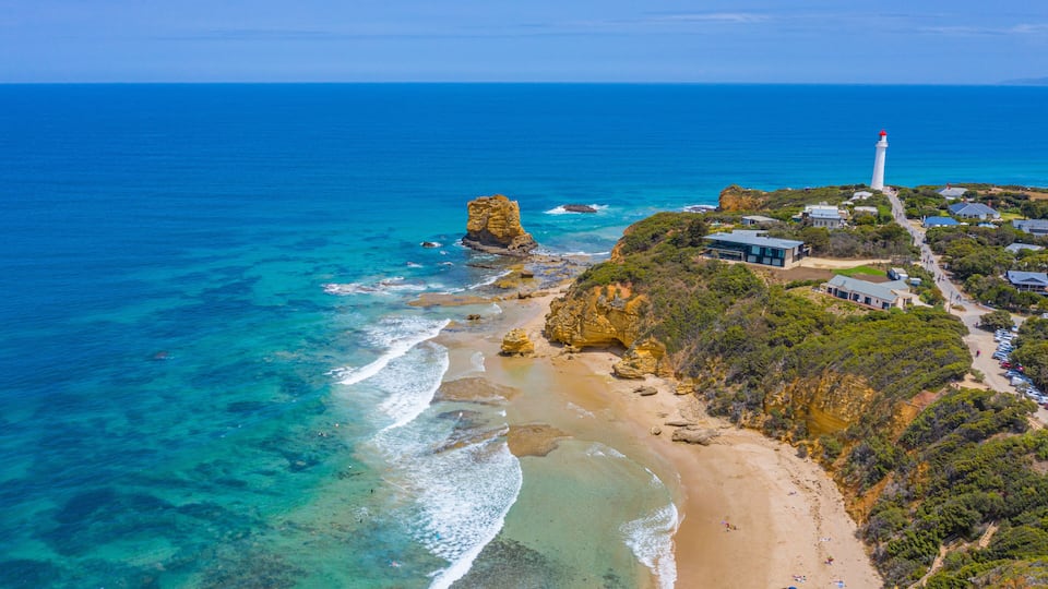 A coastal landscape with a prominent rock formation, a lighthouse, and a beach with clear waters.