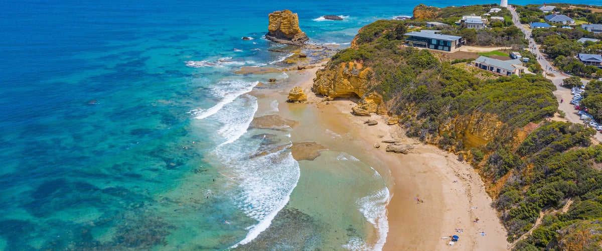 A coastal landscape with a prominent rock formation, a lighthouse, and a beach with clear waters.