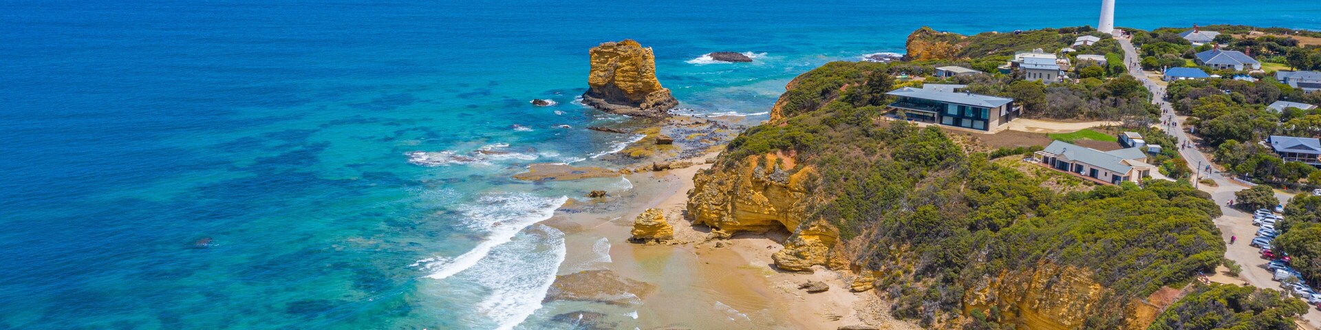 A coastal landscape with a prominent rock formation, a lighthouse, and a beach with clear waters.