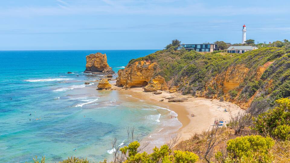A coastal landscape with a prominent rock formation, a sandy beach, and a lighthouse.