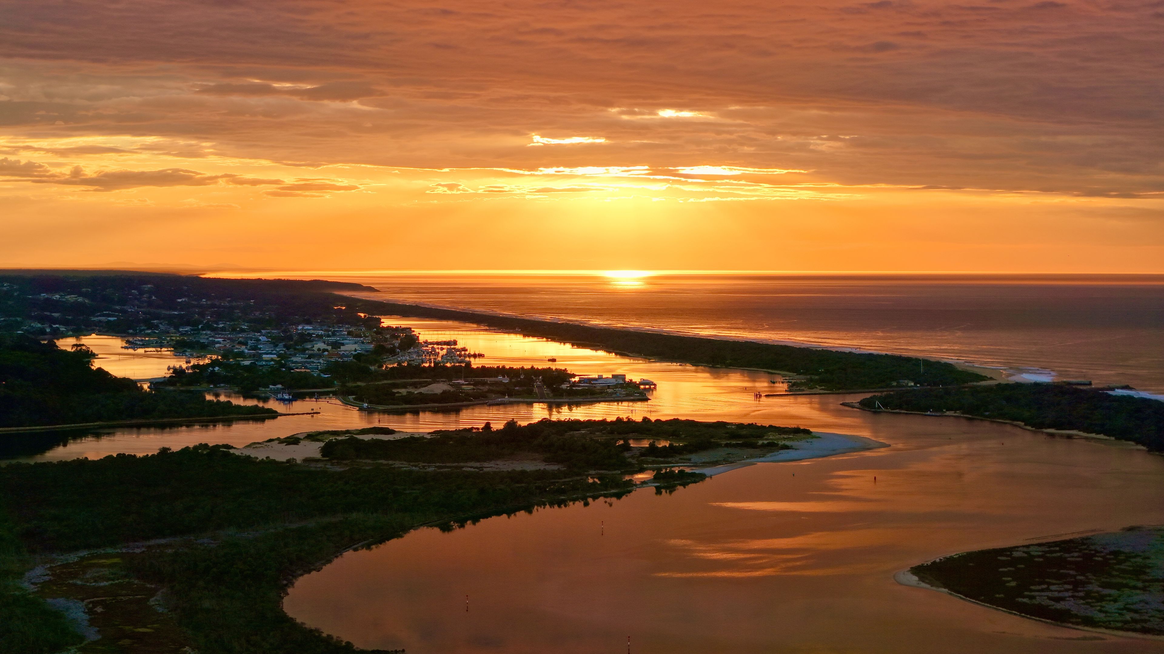 A sunset over a coastal town with a river, boats, and a bridge.