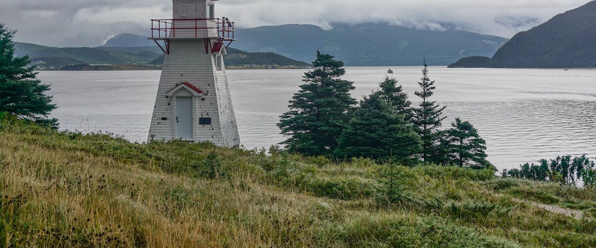 A white lighthouse with a red railing, surrounded by a grassy area and trees, overlooking a calm body of water with mountains in the background.