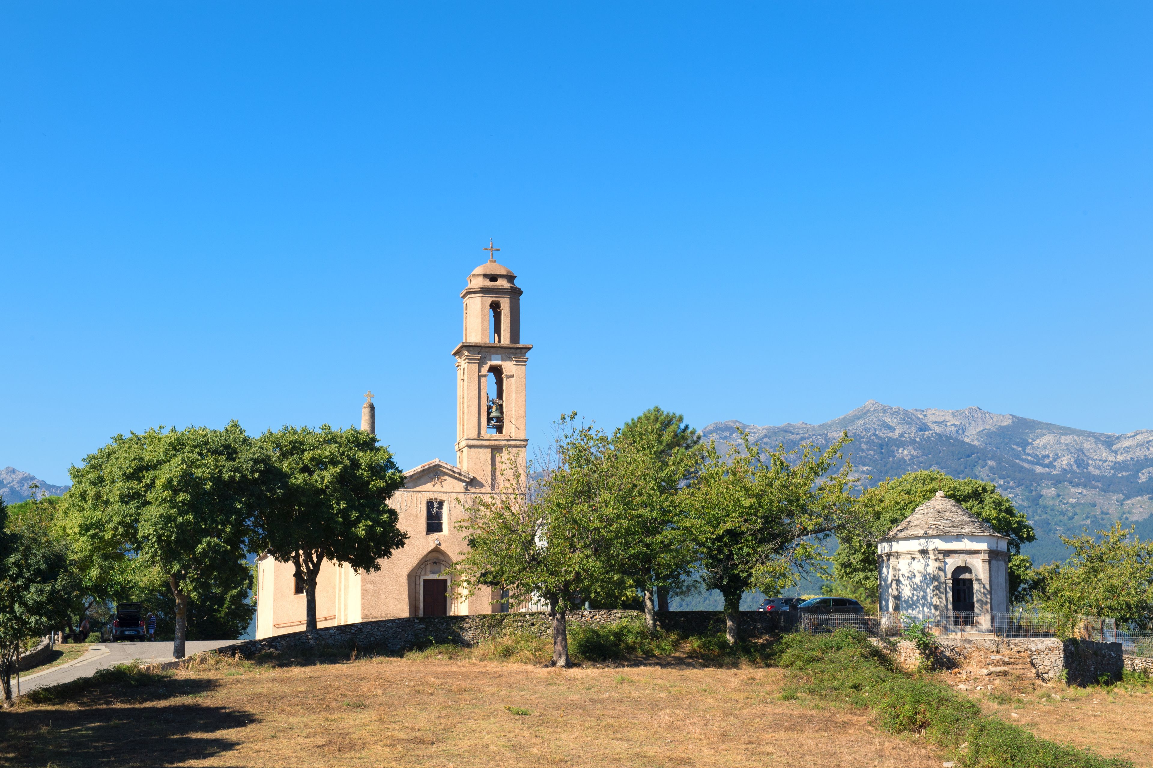 Eine Kirche mit einem Glockenturm, umgeben von Bäumen und Bergen im Hintergrund.