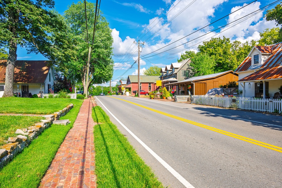 A sunny street lined with houses, a brick sidewalk, and a white picket fence.