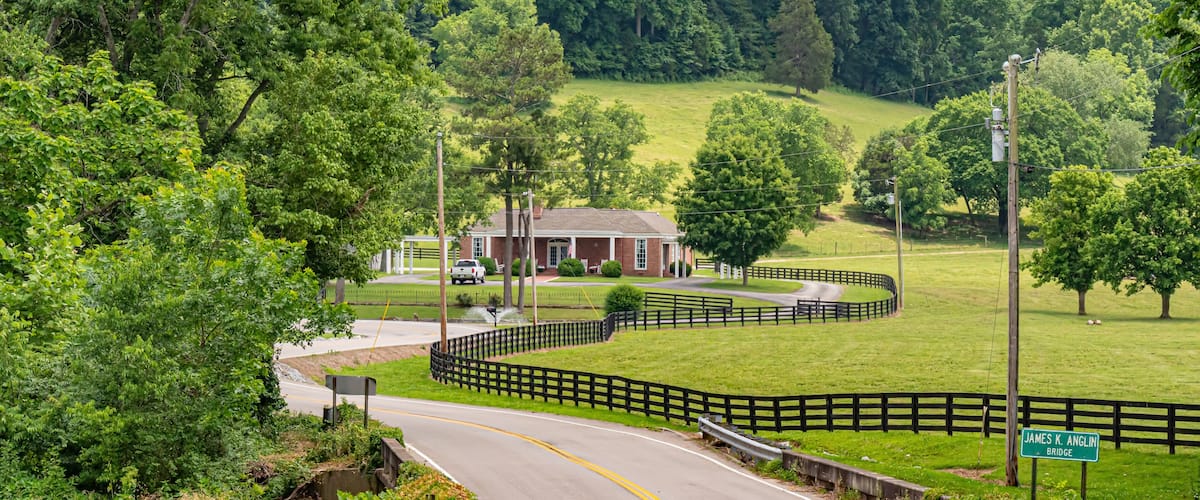 A rural road curving through a green landscape with a wooden fence, a brick building, and a sign for James K. Anglin Bridge.