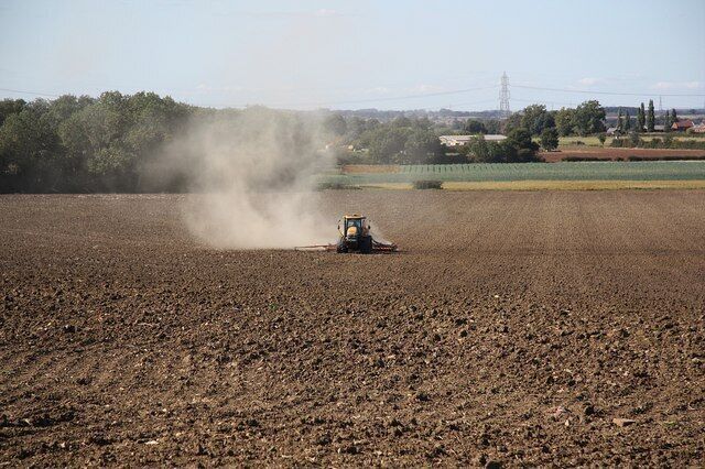 A tractor in a plowed field with dust clouds, surrounded by trees and distant buildings.