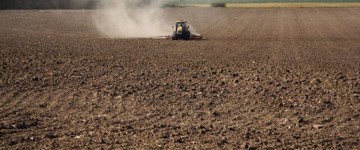 A tractor in a plowed field with dust clouds, surrounded by trees and distant buildings.