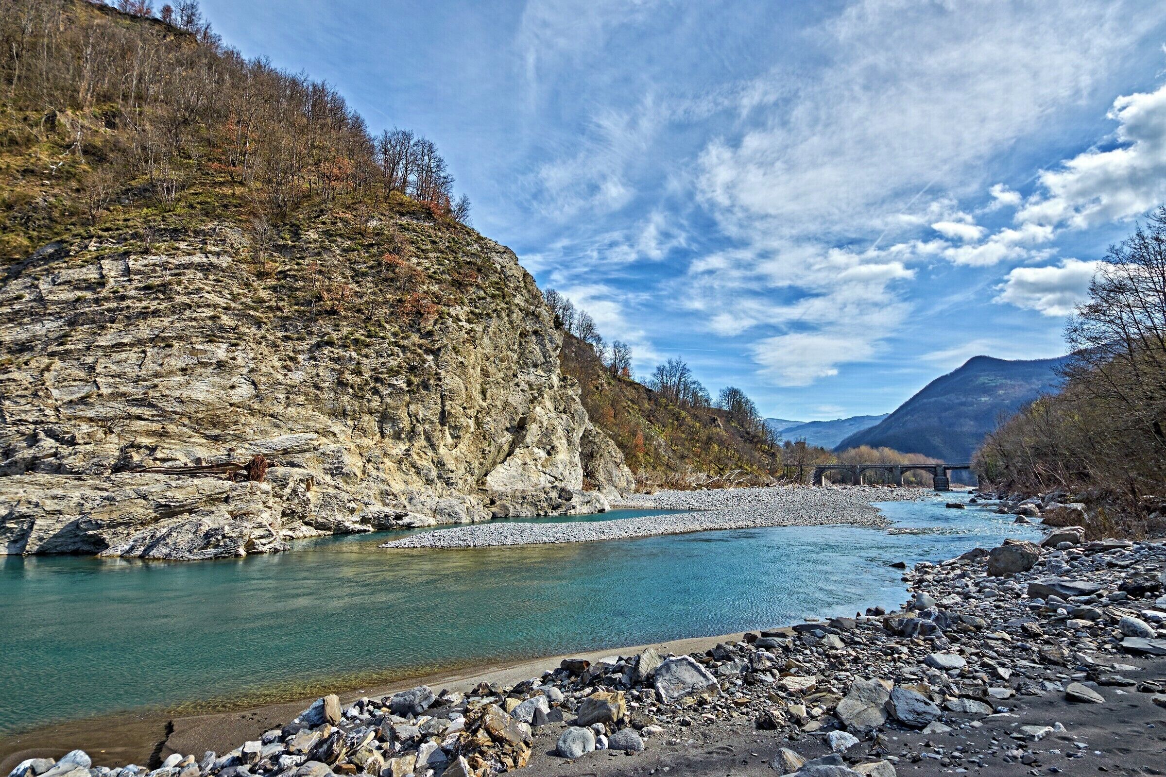 A river with rocky banks, a bridge, and mountains in the background.