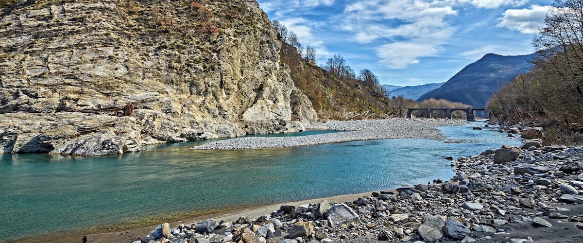 A river with rocky banks, a bridge, and mountains in the background.