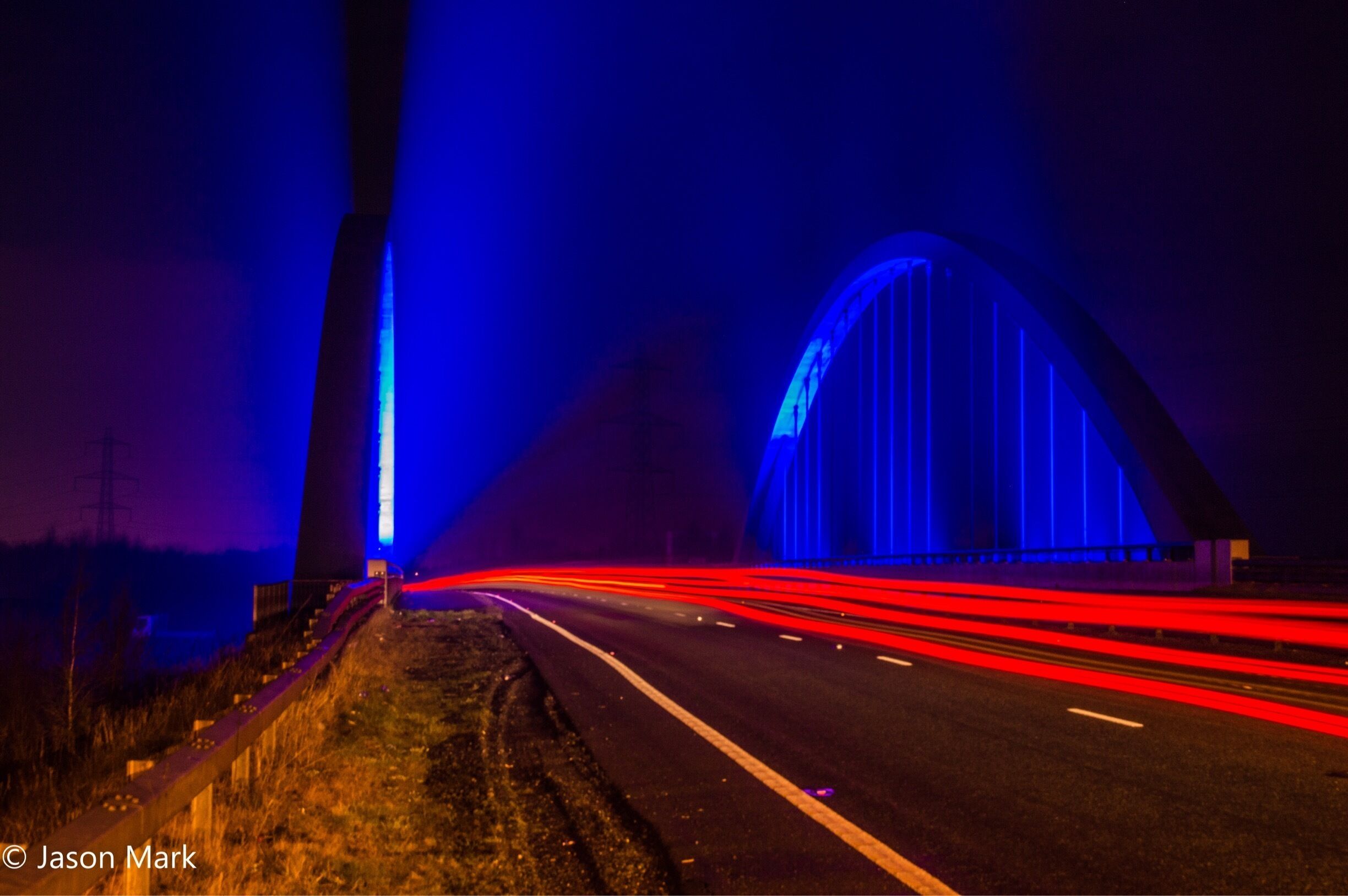 A highway at night with light trails from vehicles, a bridge with blue lighting, and a tall structure on the left.