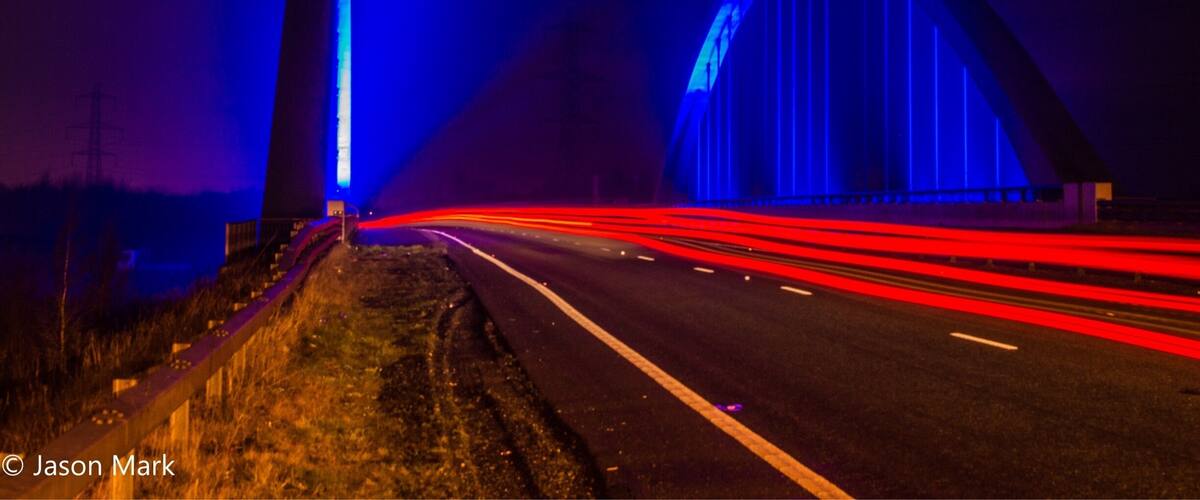 A highway at night with light trails from vehicles, a bridge with blue lighting, and a tall structure on the left.