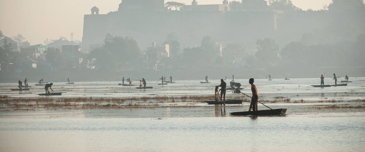A calm river with people on small boats, a distant building, and trees.