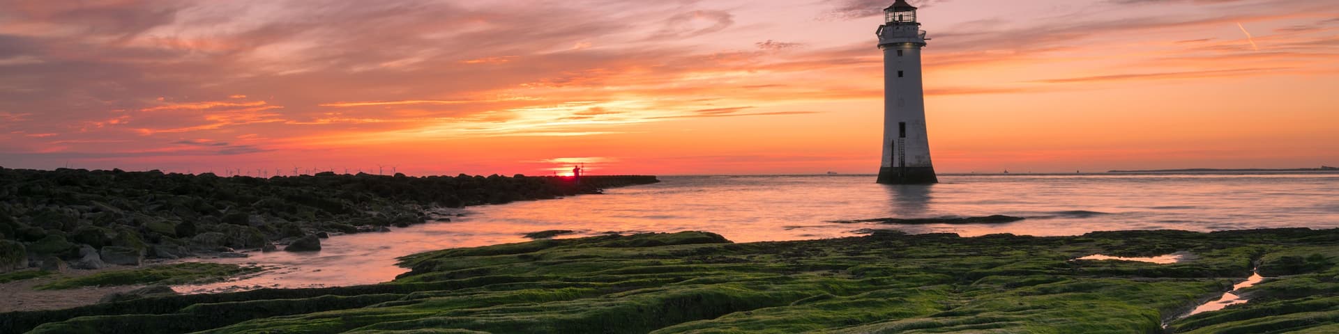 A lighthouse at sunset with a colorful sky, green moss-covered terrain, and a calm sea.