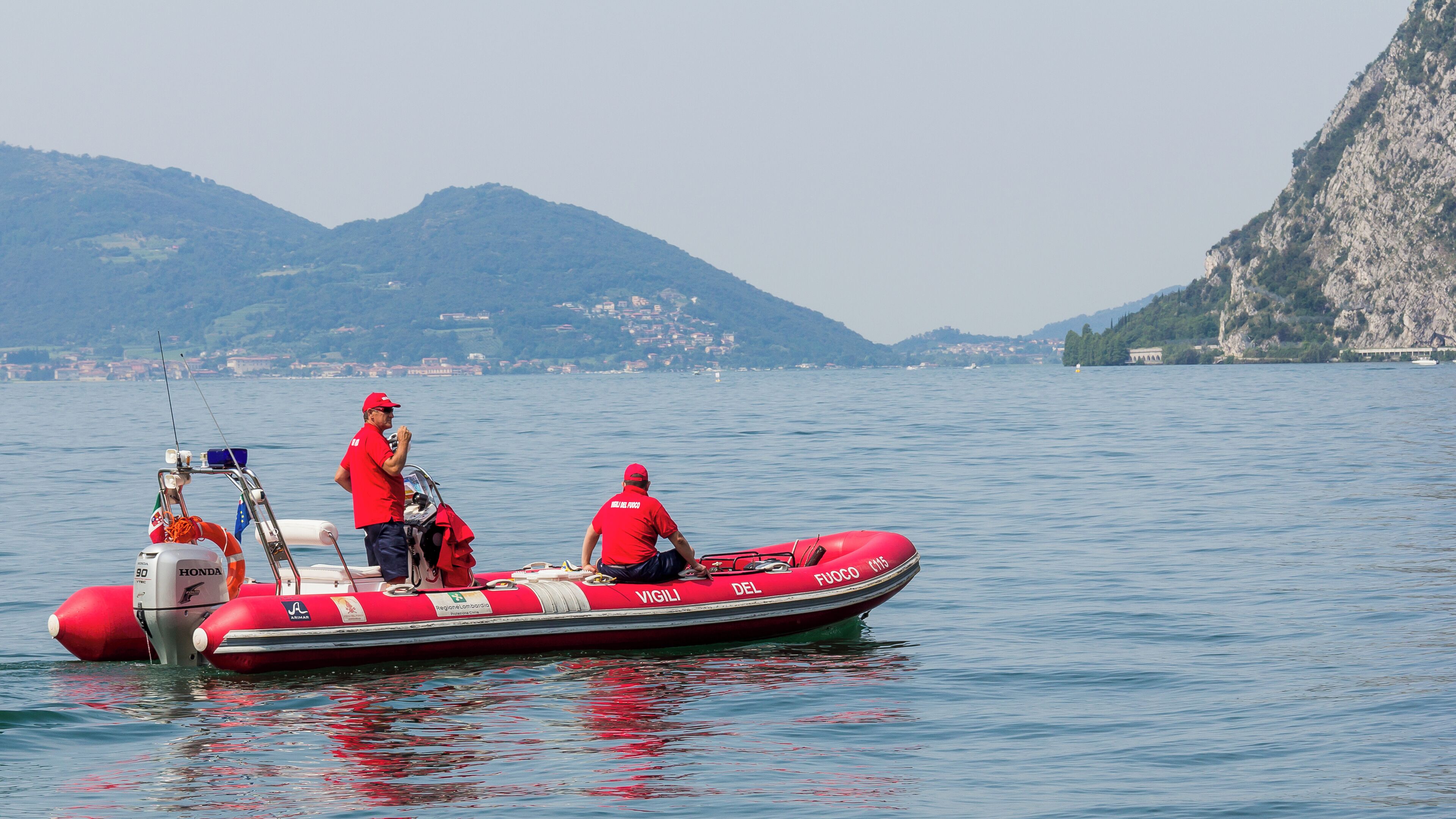 A red inflatable boat on calm water with two people in red uniforms, a mountainous landscape in the background, and a clear sky.