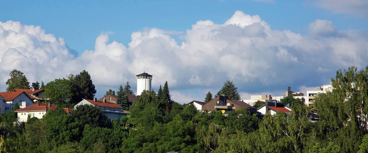 A residential area with houses, a tower, and a forested landscape under a partly cloudy sky.