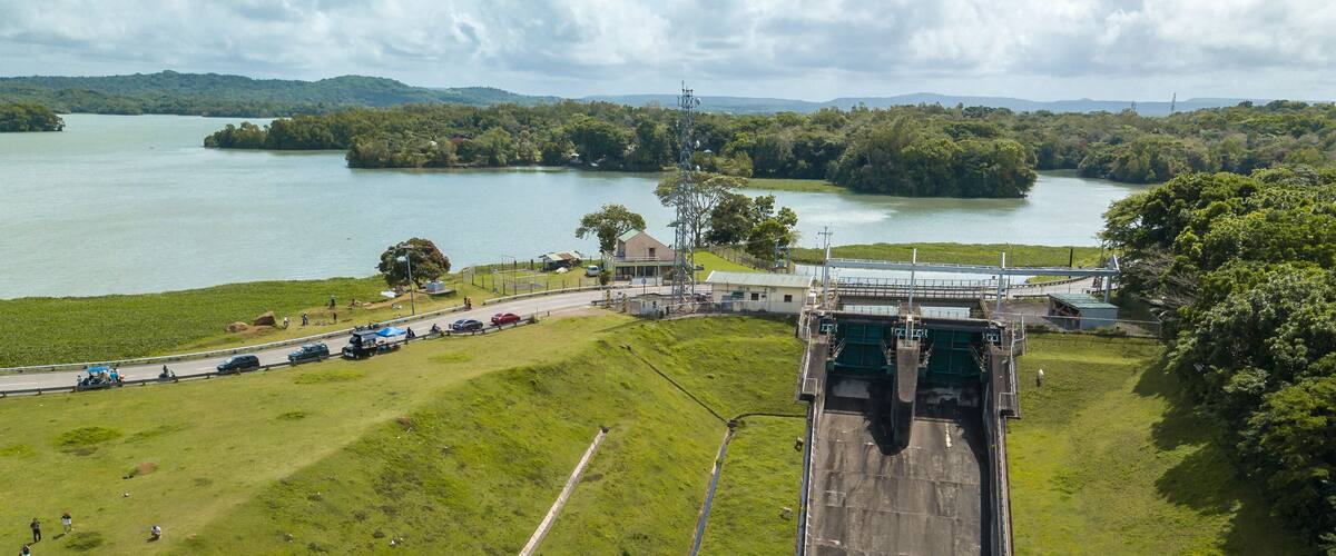 A dam with a spillway, a road with vehicles, and a lake surrounded by greenery.
