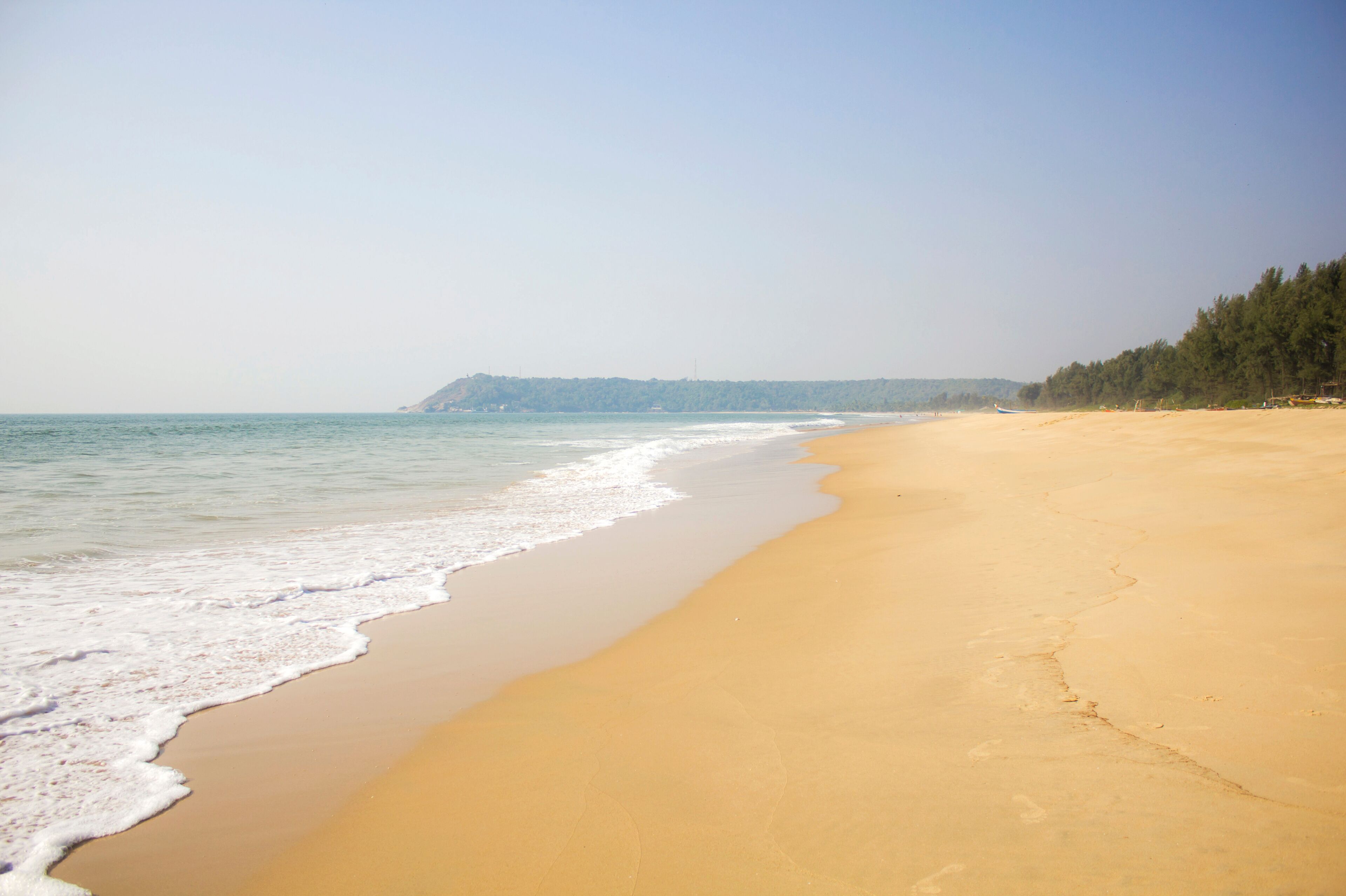 A sandy beach with gentle waves, a clear sky, and a forested hill in the background.