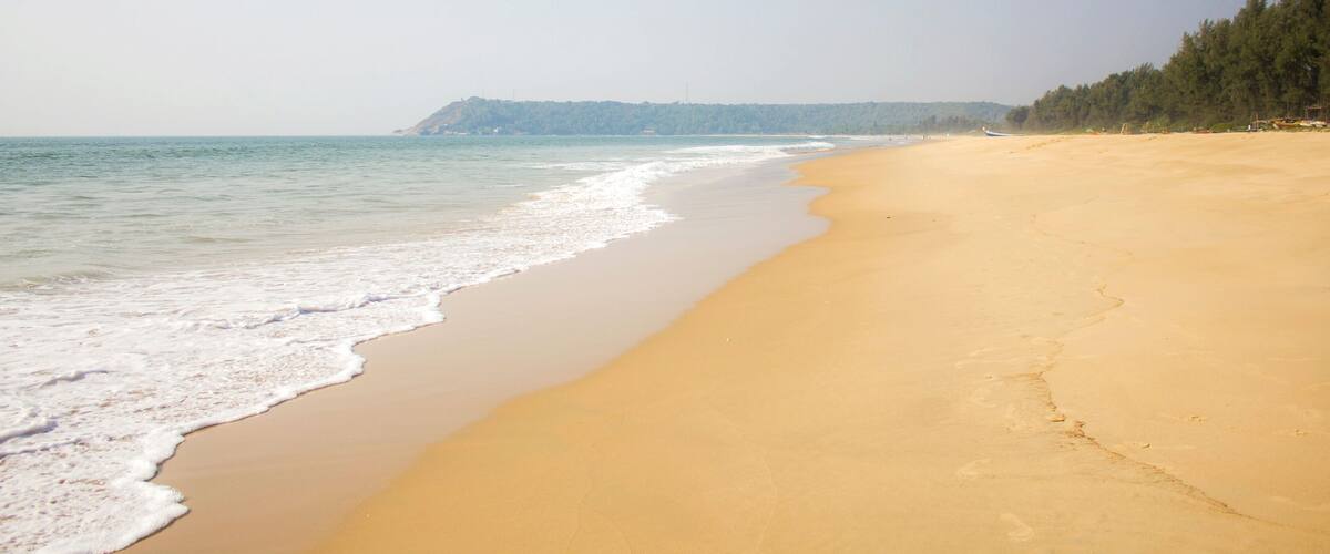 A sandy beach with gentle waves, a clear sky, and a forested hill in the background.