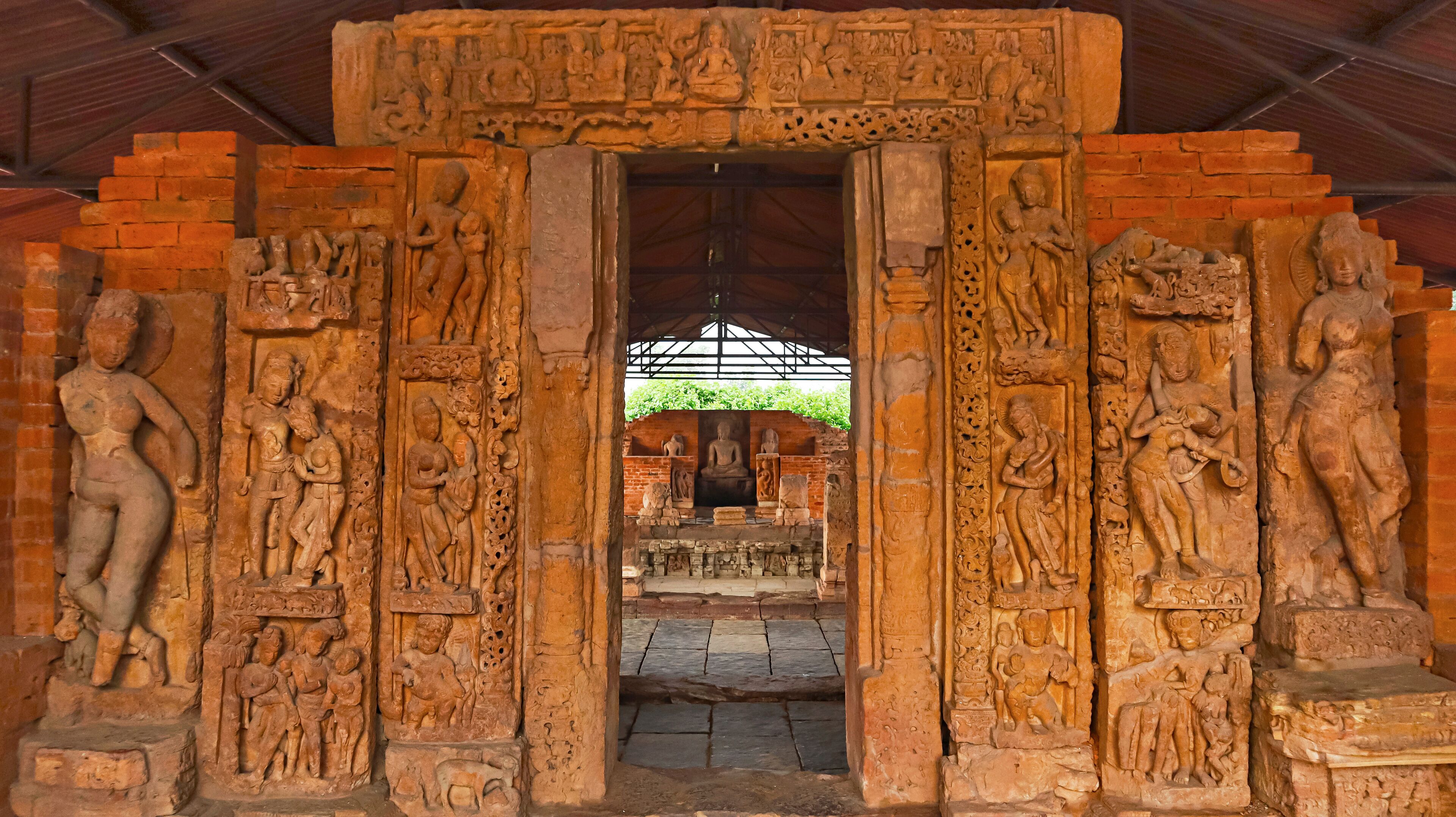 A stone doorway with intricate carvings of figures and deities, leading to a covered area with a Buddha statue.