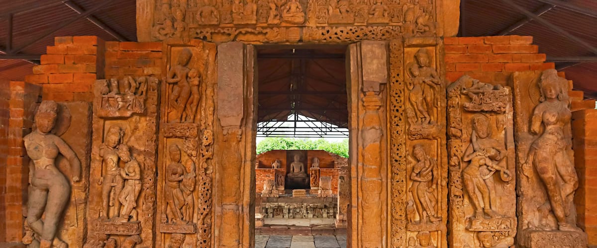 A stone doorway with intricate carvings of figures and deities, leading to a covered area with a Buddha statue.
