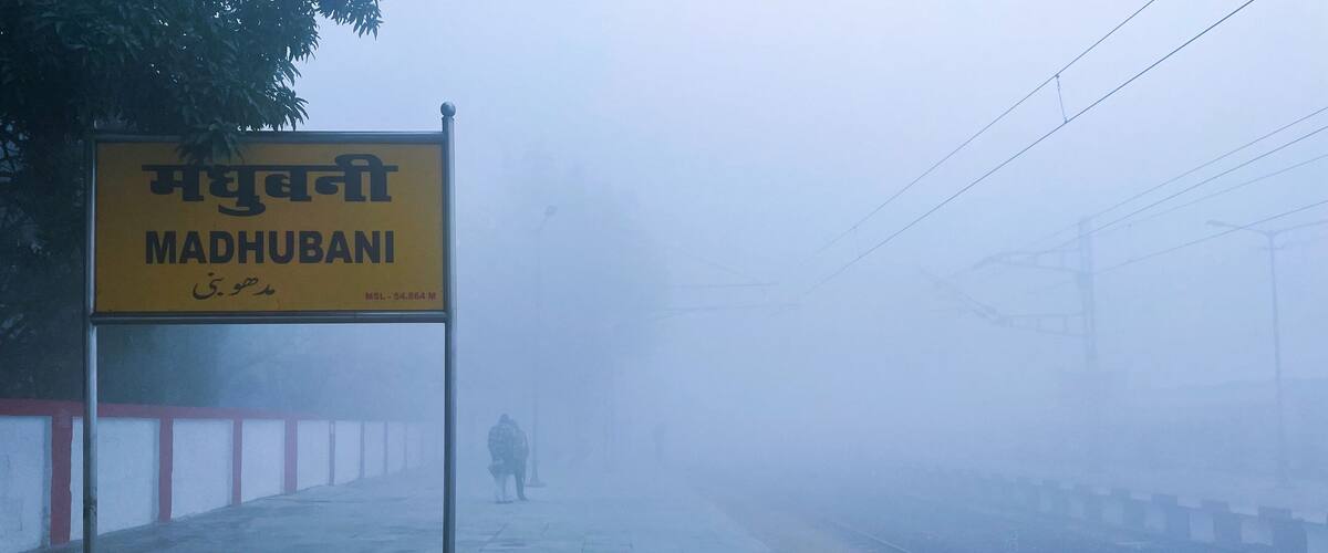 A foggy train station platform with a sign reading "Madhubani" and a person walking in the distance.