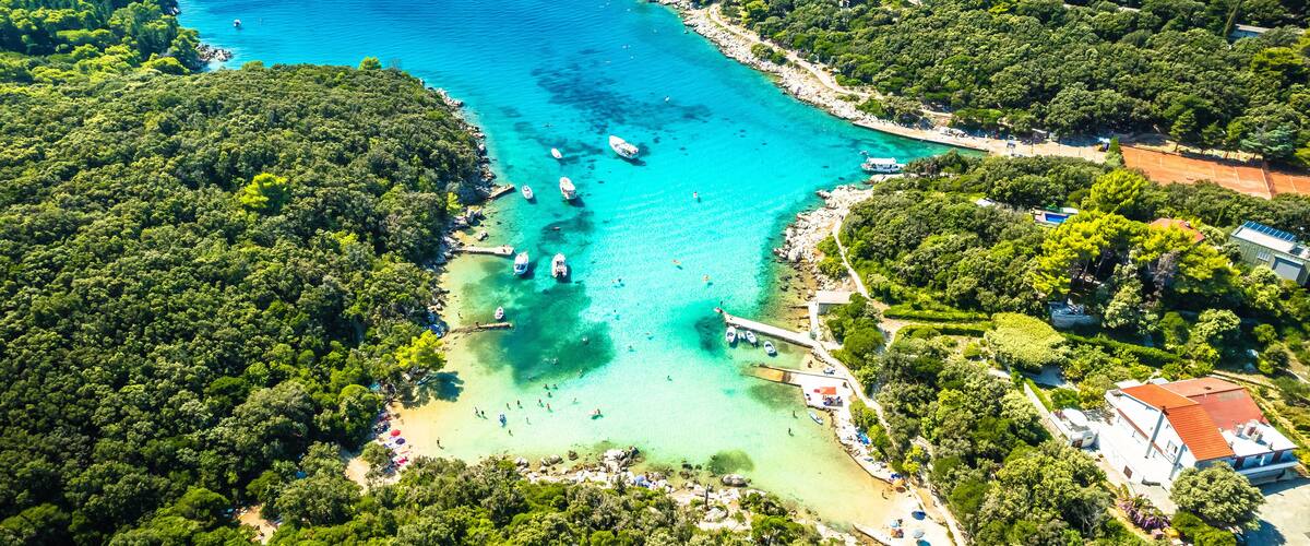 An aerial view of a coastal area with clear turquoise waters, a small beach, and dense green forests.